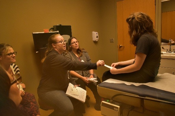 Five Caucasian women in a patient room observing two Caucasian women demonstrate